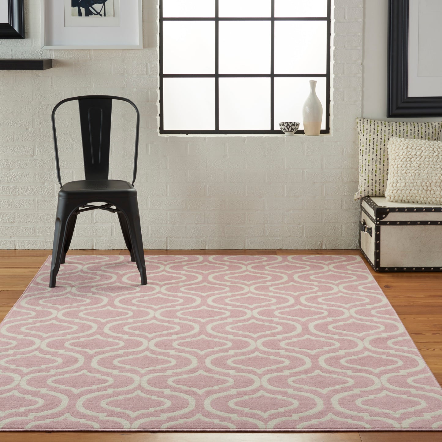 Black metal chair on a pink wavy-pattern rug in a bright white brick-walled living room with window, beige sofa & pillows.