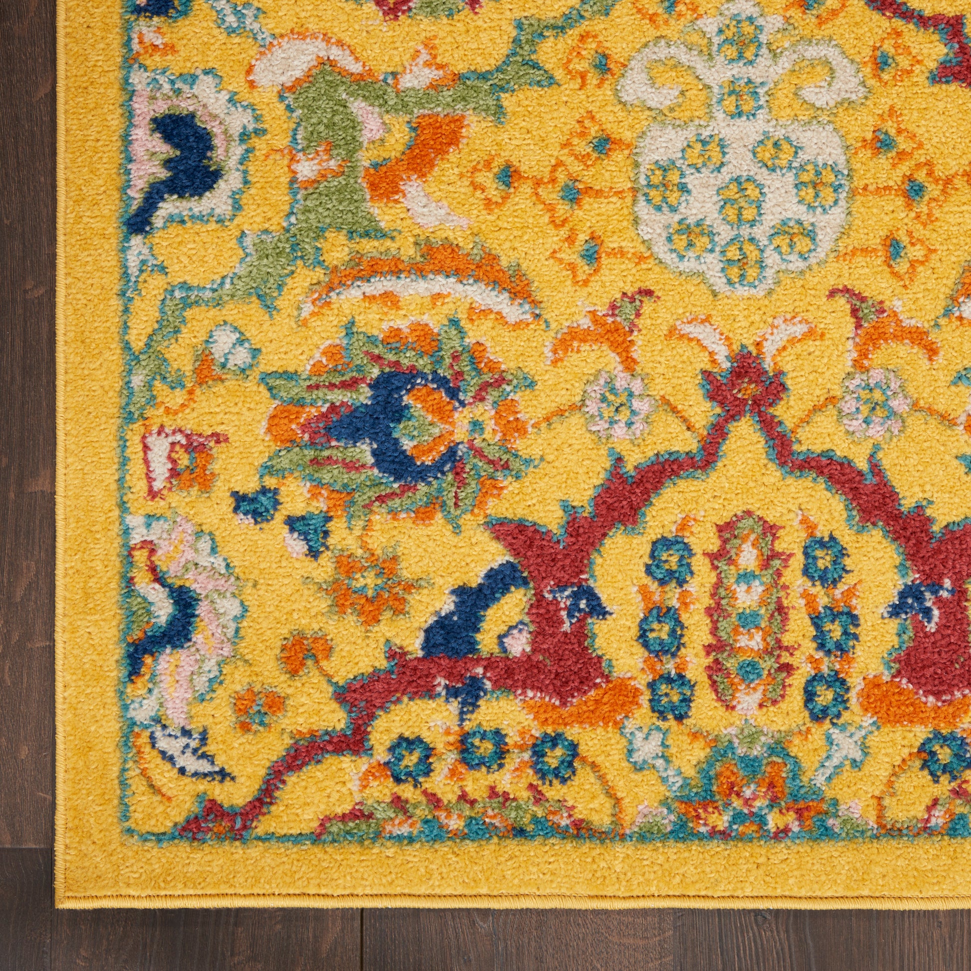 Corner of a vibrant yellow oriental rug with blue, red, teal floral motifs & a gold border on a dark wooden floor.