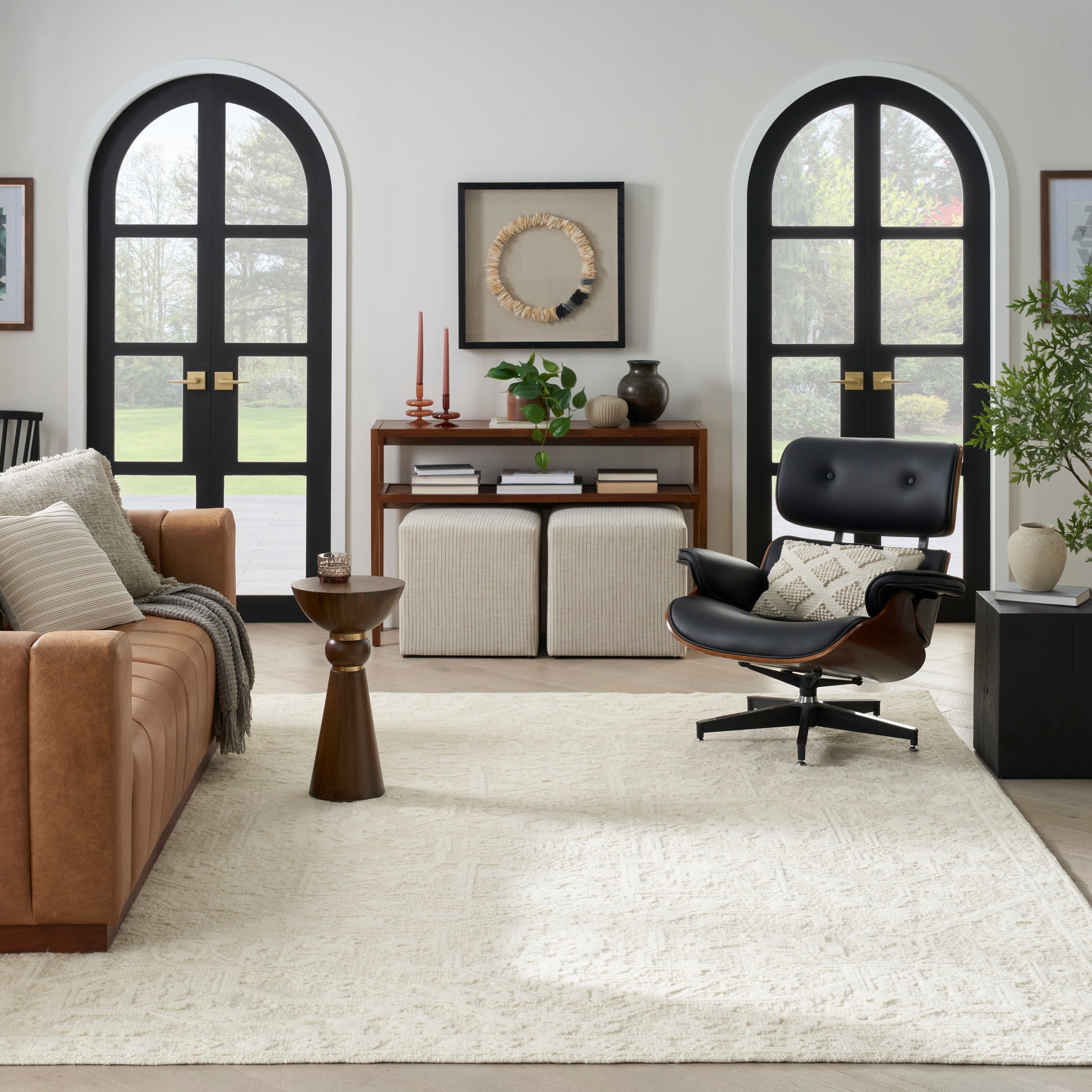 Living room with tan leather sofa, black Eames lounge chair, arched black windows, cream rug, and wooden console with plants.