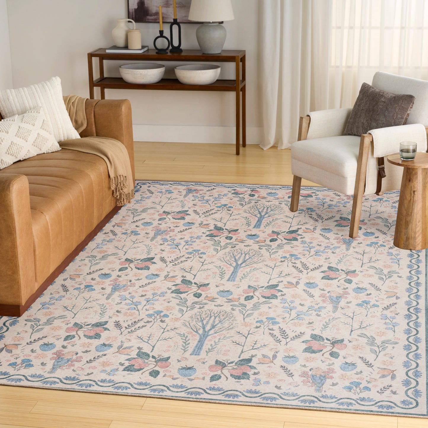 Cream area rug with blue botanical motif and border in a sunny living room with a tan leather sofa and white armchair.