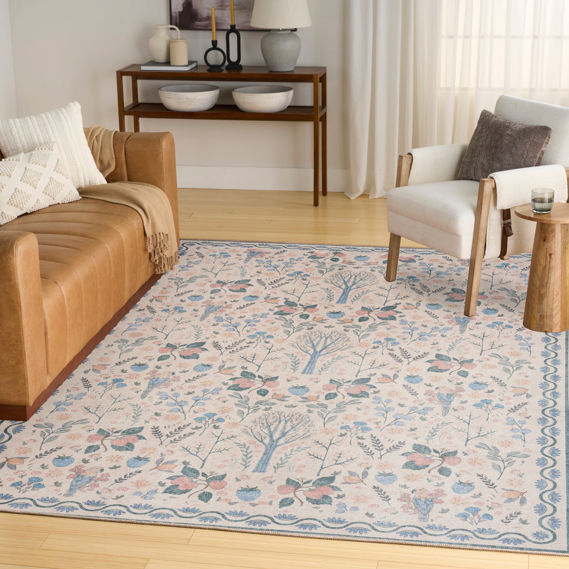 Cream area rug with blue botanical motif and border in a sunny living room with a tan leather sofa and white armchair.