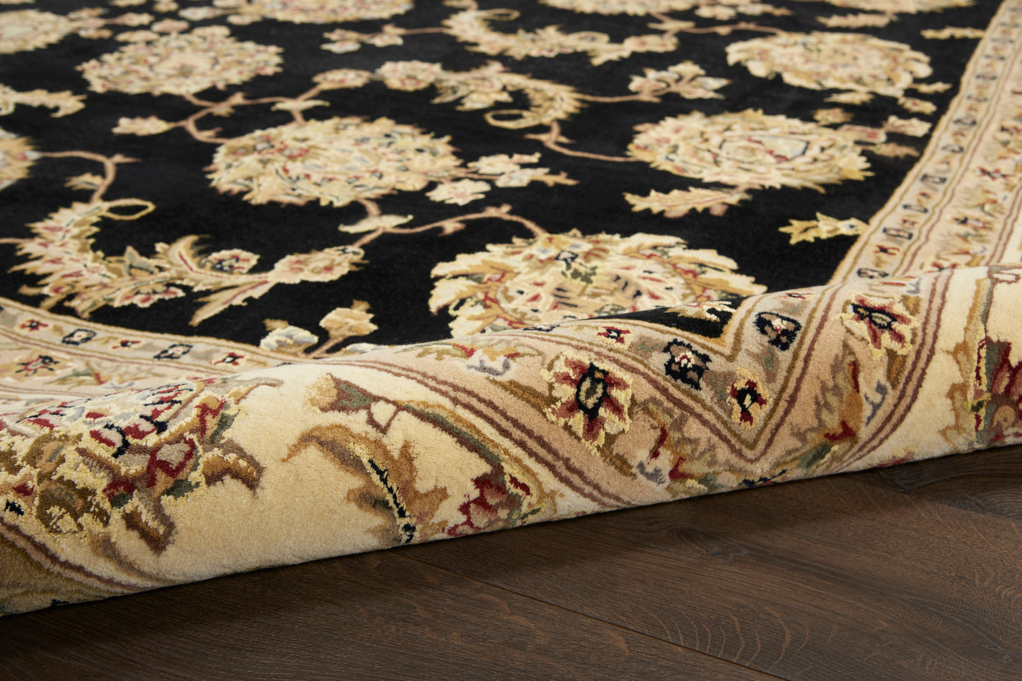 Black oriental rug with cream floral border, gold and burgundy accents, rolled edge on dark wood floor.