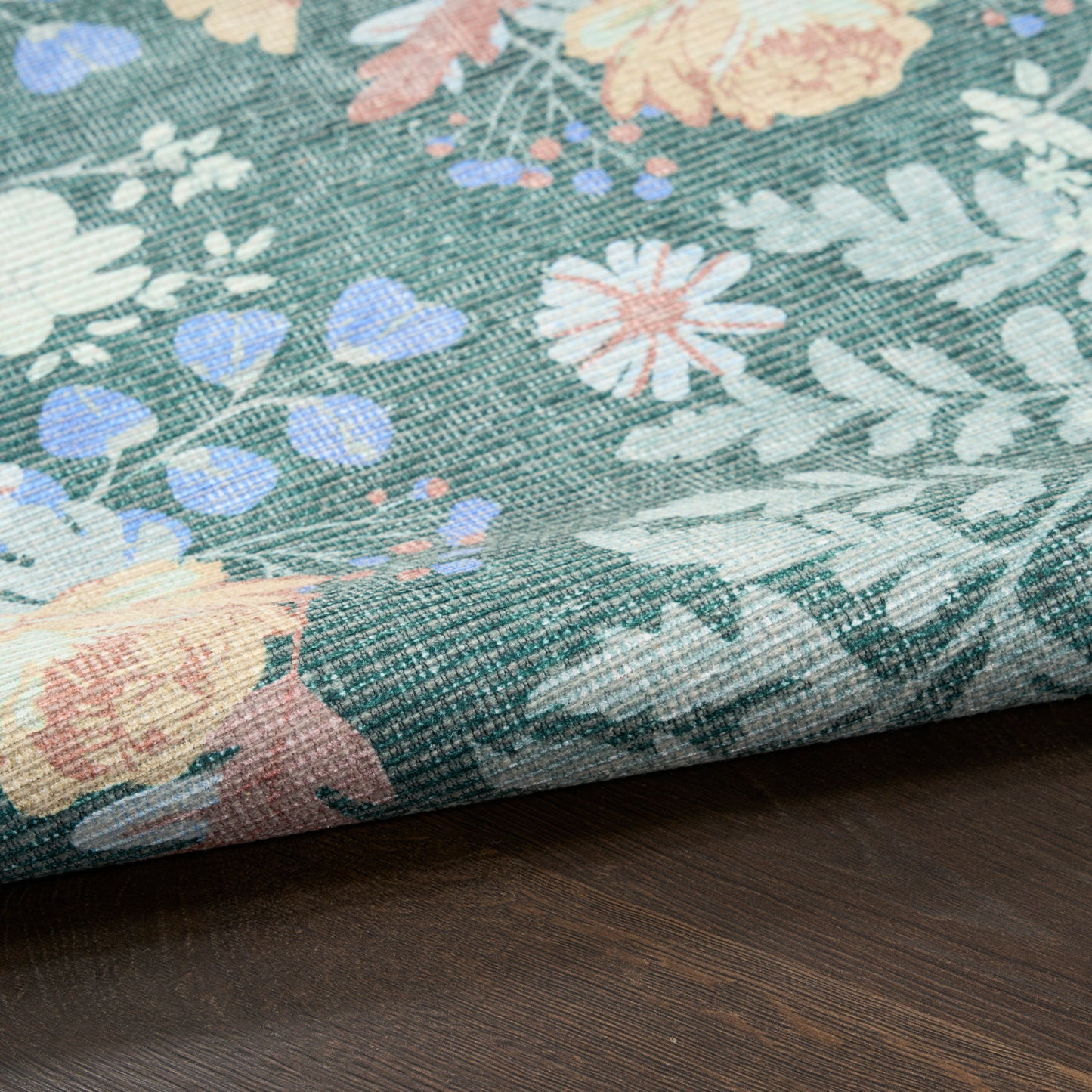 Floral rug with teal background & pastel pink, peach, white, and blue flowers; folded edge visible on dark wood surface; 4' x 6'.