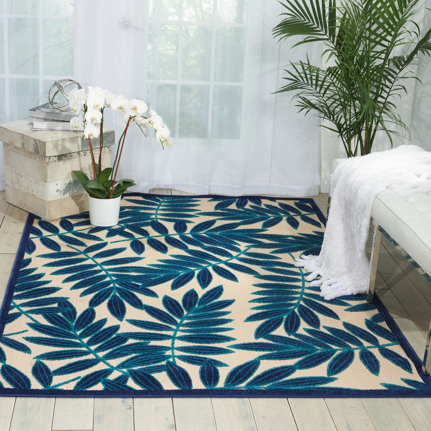 Beige area rug with navy blue tropical leaf pattern, laid in a bright room with white orchid, palm plant & white throw.