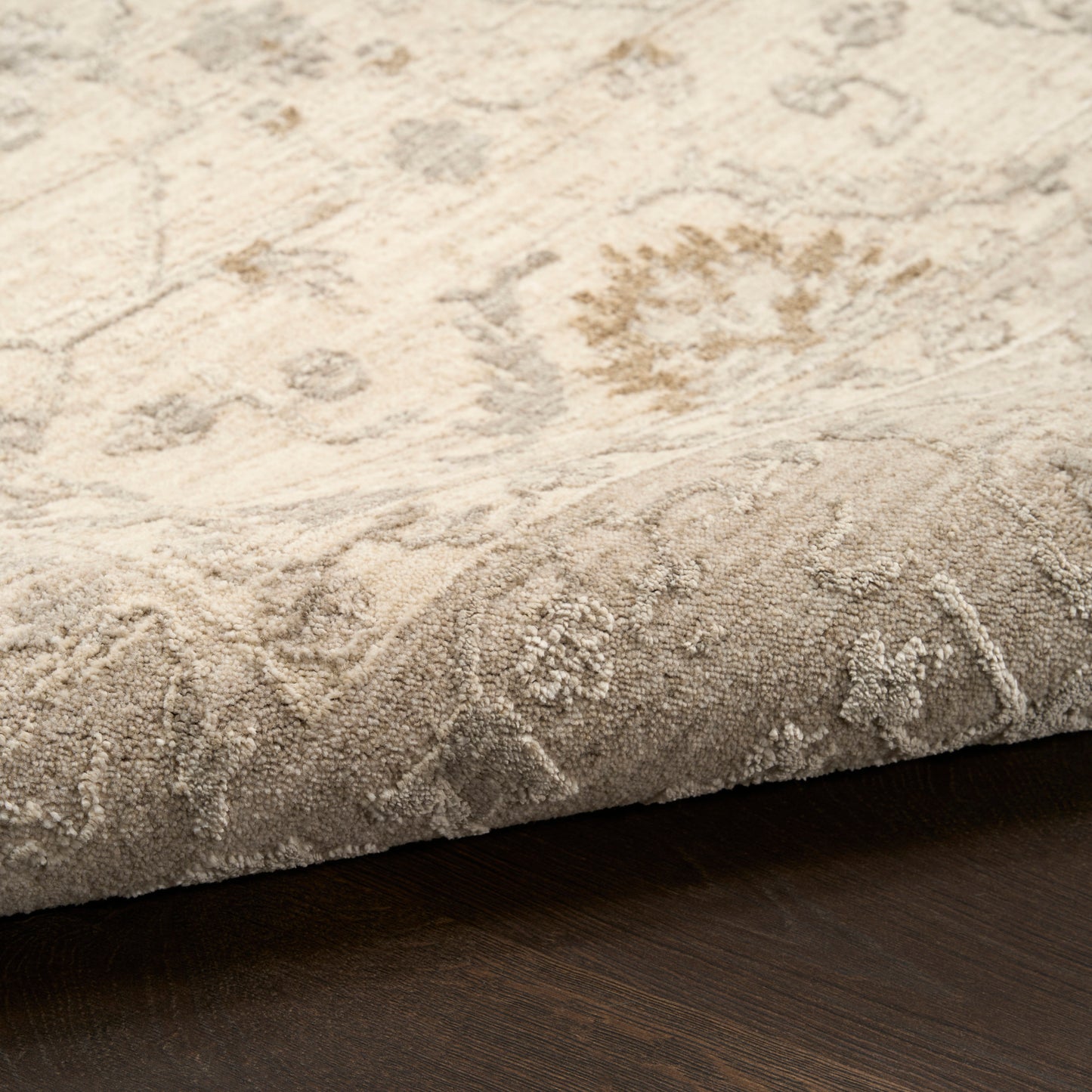 Beige floral patterned area rug with plush pile; rolled edge shows thickness on dark wood floor.