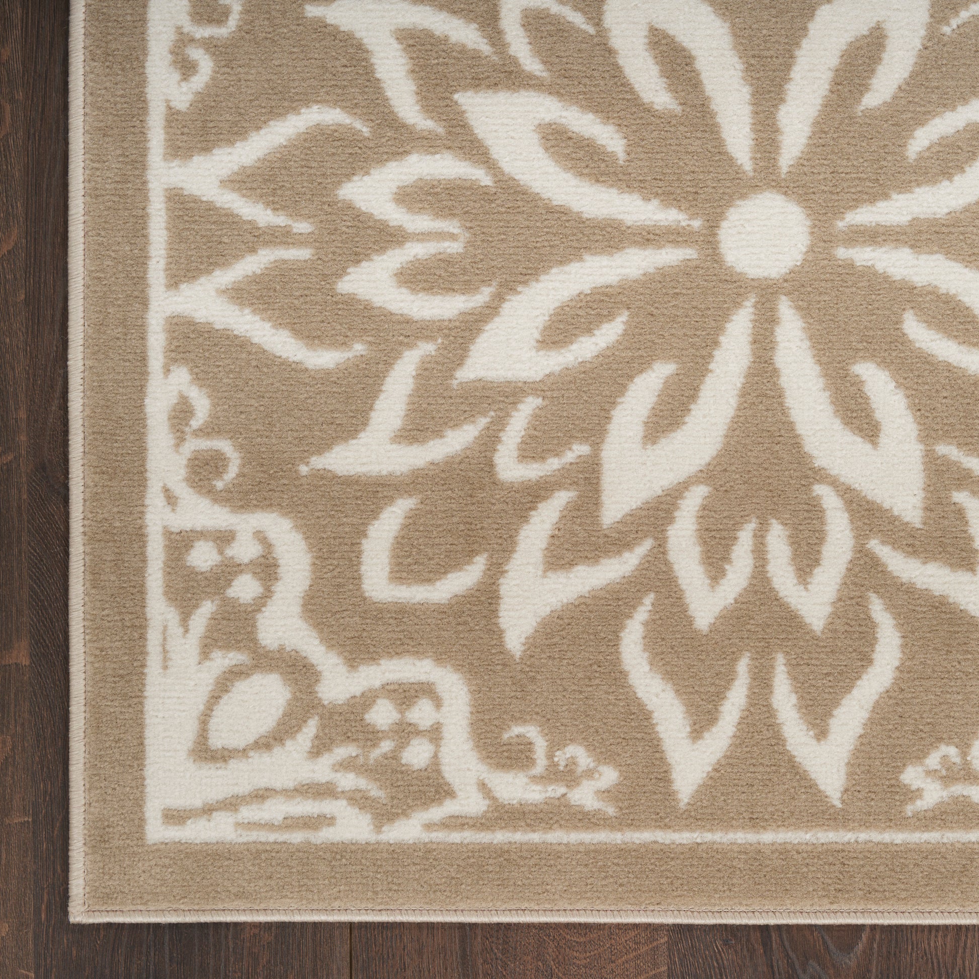 Beige area rug with white floral medallion pattern and border on dark wood floor.