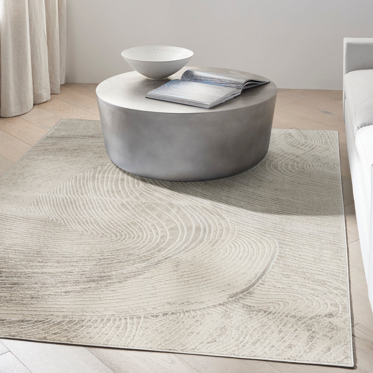Round concrete-look gray coffee table with a white bowl and open magazine on a beige patterned rug.