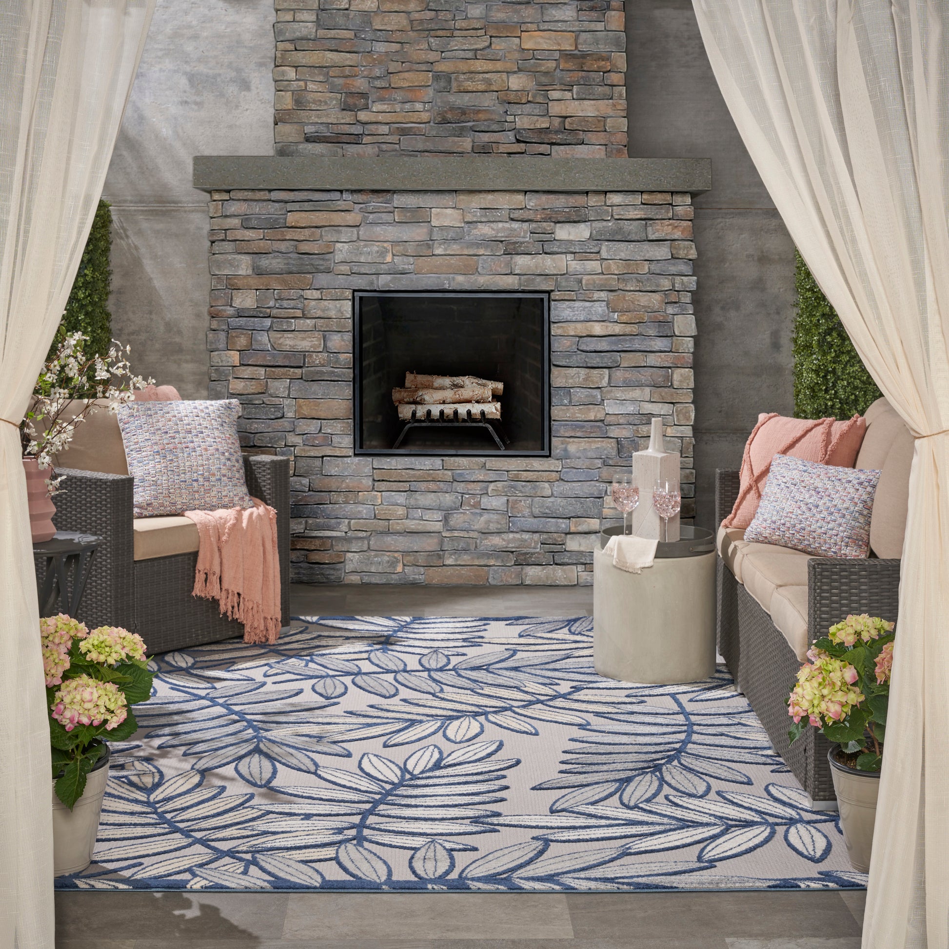 Outdoor lounge with stone fireplace centerpiece, wicker sofas, blue leaf rug, cream curtains, and potted flowers.