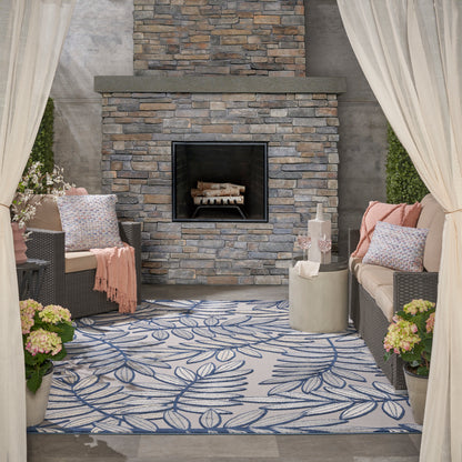 Outdoor lounge with stone fireplace centerpiece, wicker sofas, blue leaf rug, cream curtains, and potted flowers.