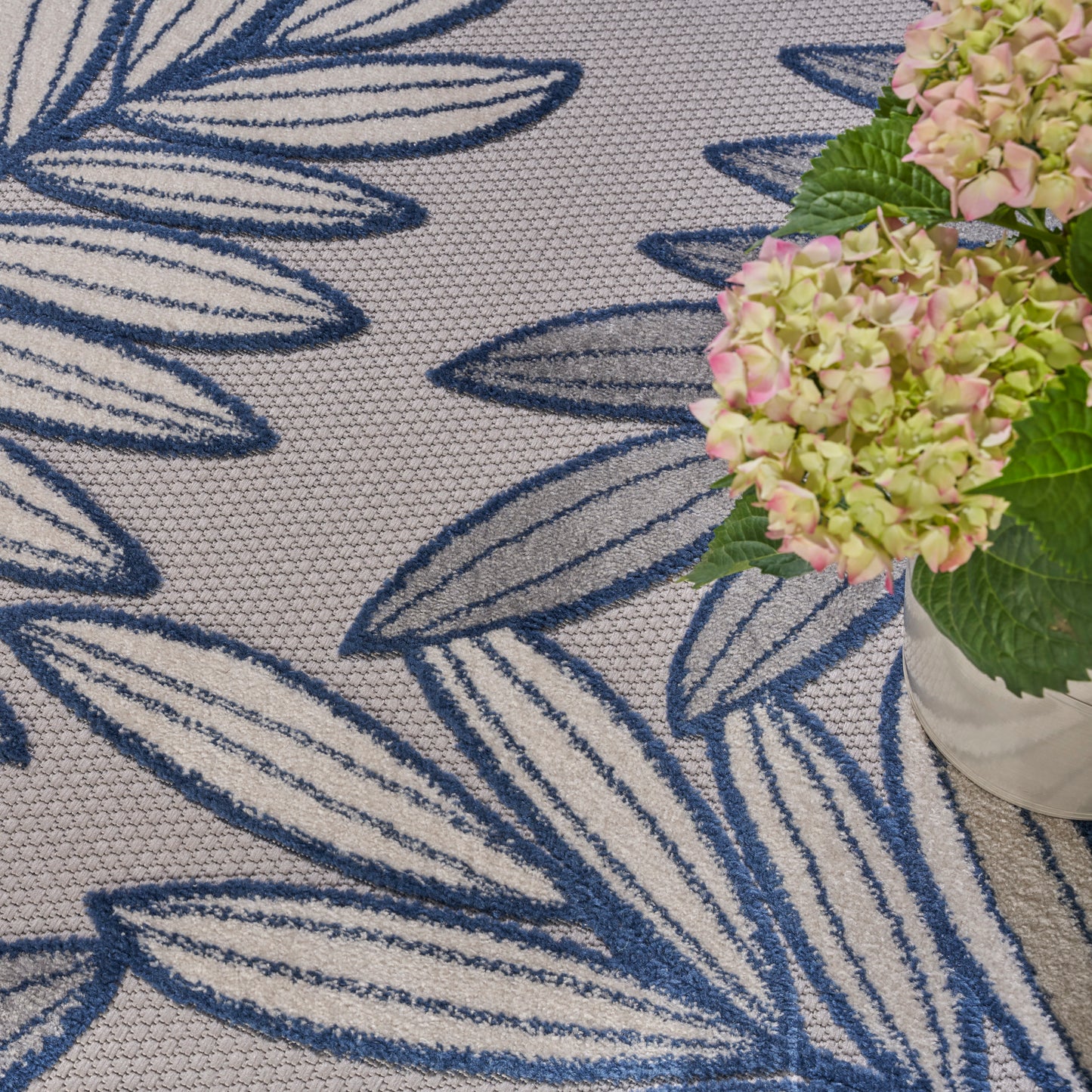Beige woven rug with bold blue leaf pattern, beside a pink hydrangea bouquet in a white vase.