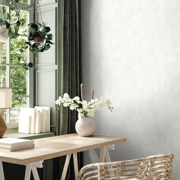 Wallpaper with a light industrial white design on the wall behind a light wood desk; visible items include a white orchid vase, stacked books, a hanging plant, green drapes, a sunlit window, and a woven wicker chair.