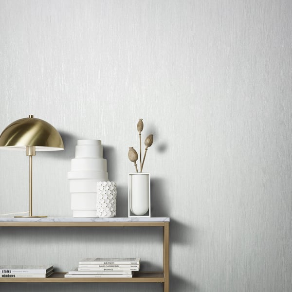 White wallpaper with subtle air-slub texture as backdrop; foreground shows a gold-dome lamp and white ceramic decor on a gold-framed shelf: stacked discs, a honeycomb vase, and a tall vase with dried pods.