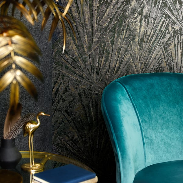 Wallpaper featuring teal & gold palm leaves; foreground shows a teal velvet chair beside a round gold table with blue books, a gold flamingo statue, & a black vase.