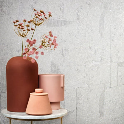 Beige wallpaper with a graphic, airy pattern; on a round marble table sits a tall rust-colored vase with dried pink flowers and two pink ceramic pots.