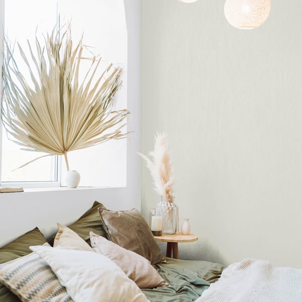 White textured wallpaper on the wall of a boho bedroom; visible decor includes a dried palm by the window, beige-green pillows, woven textiles, and a wooden side table.