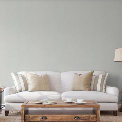 Neutral living room with light grey wallpaper featuring a subtle silver-grey stripe; beige and striped pillows on a light grey fabric sofa; wooden coffee table with cups and a book on a rug.