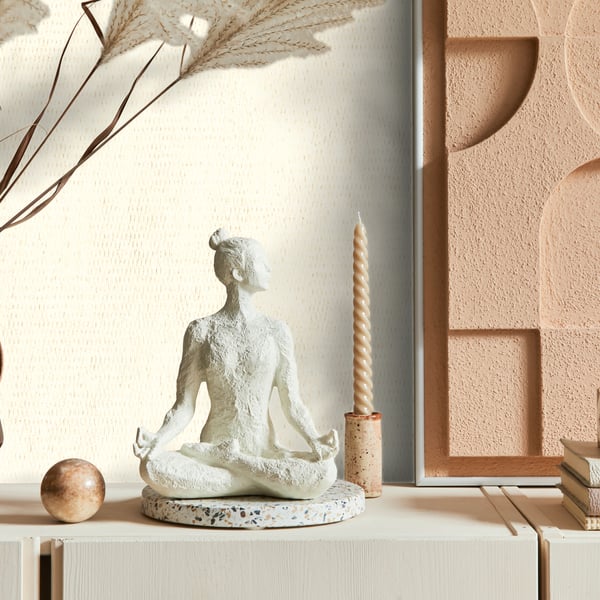 White wallpaper with subtle texture; scene includes a white seated meditation statue on a speckled base, beige twisted candle, pampas stems, peach geometric wall art & books.