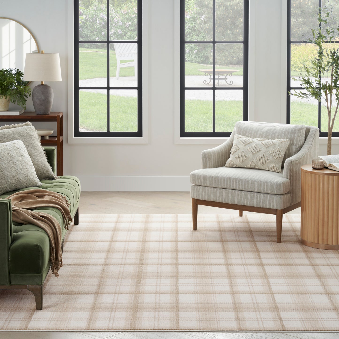 Sunlit living room with green velvet sofa, light gray striped chair, beige plaid rug, wooden side table & potted plants.