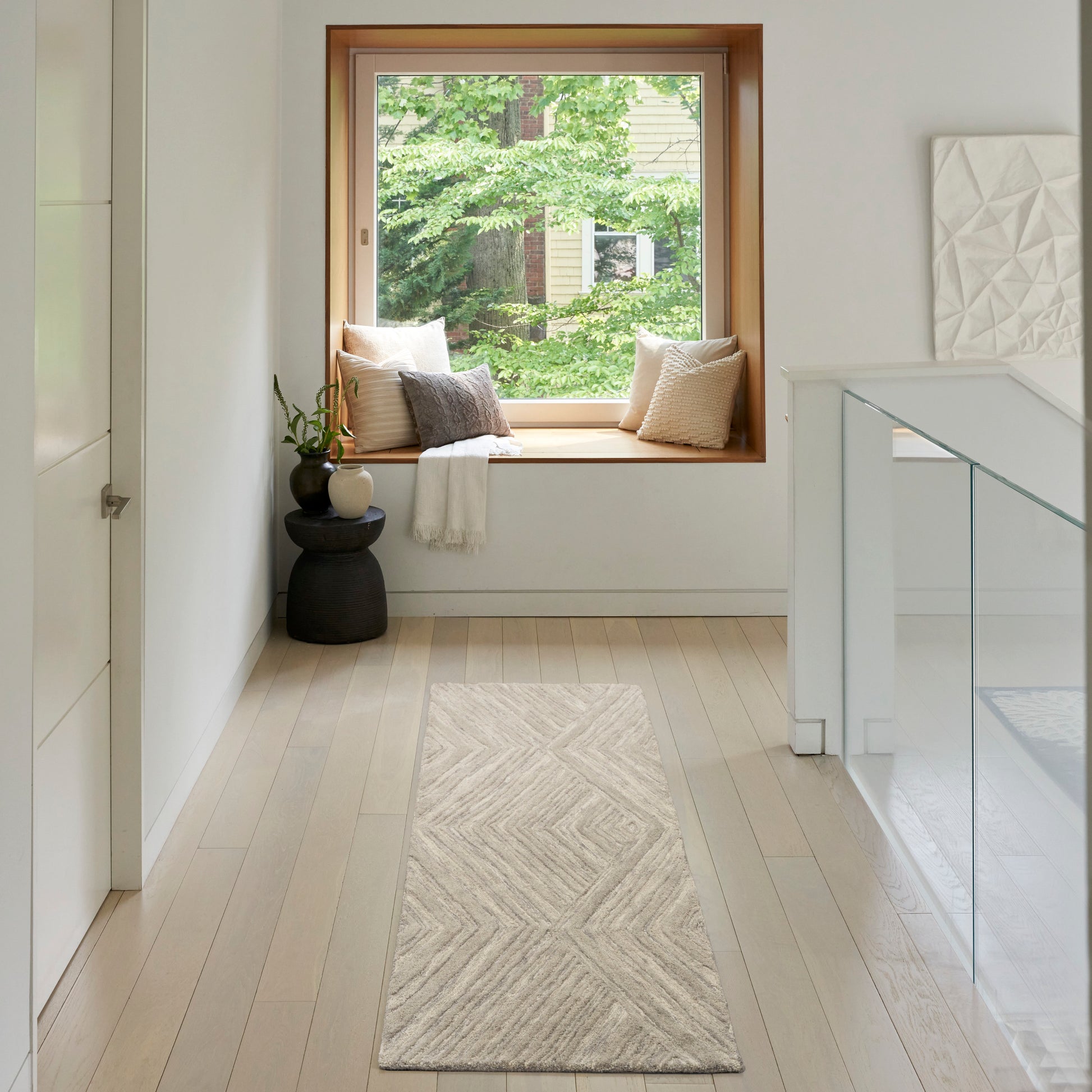 Bright hallway with light wood floors, beige chevron rug, window seat with pillows, plant, glass-railed staircase.