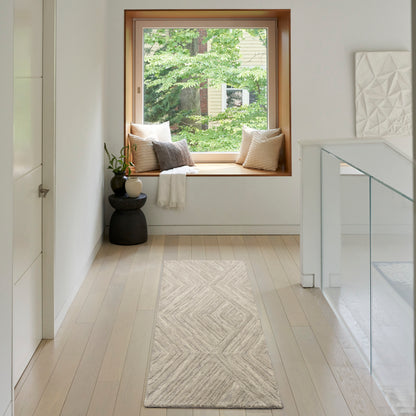 Bright hallway with light wood floors, beige chevron rug, window seat with pillows, plant, glass-railed staircase.