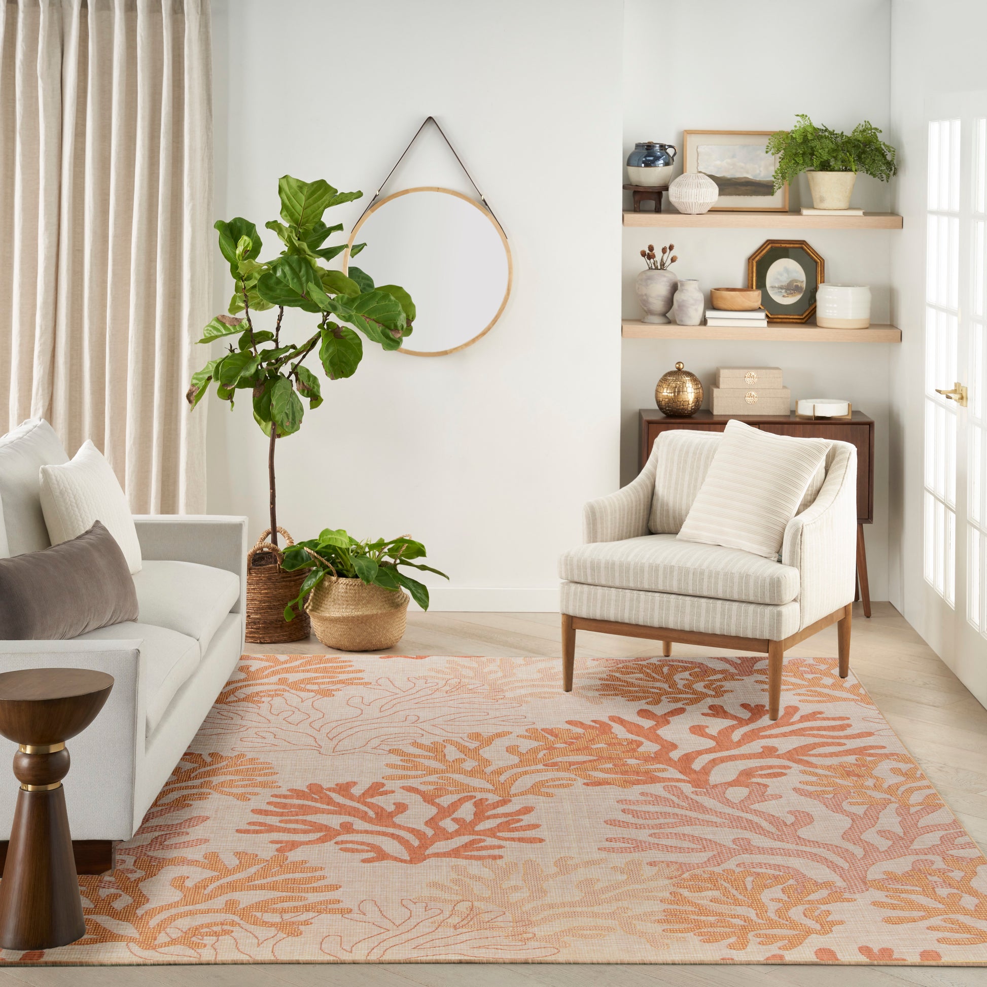 Cozy living room with orange coral rug, cream armchair and sofa, large fiddle-leaf fig, round wall mirror, wooden shelves