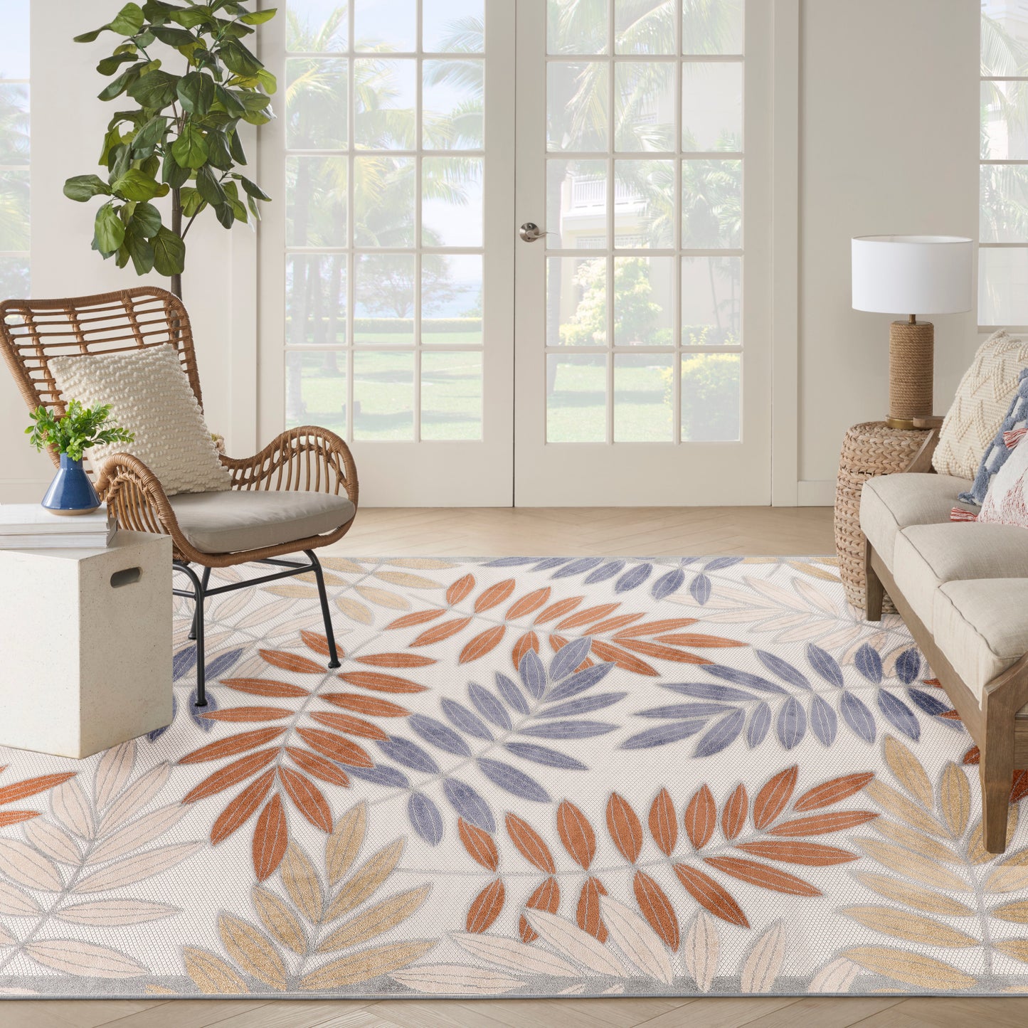 Bright outdoor space with a leaf-pattern rug in orange, blue, and beige, a rattan chair, plants, and glass patio doors.