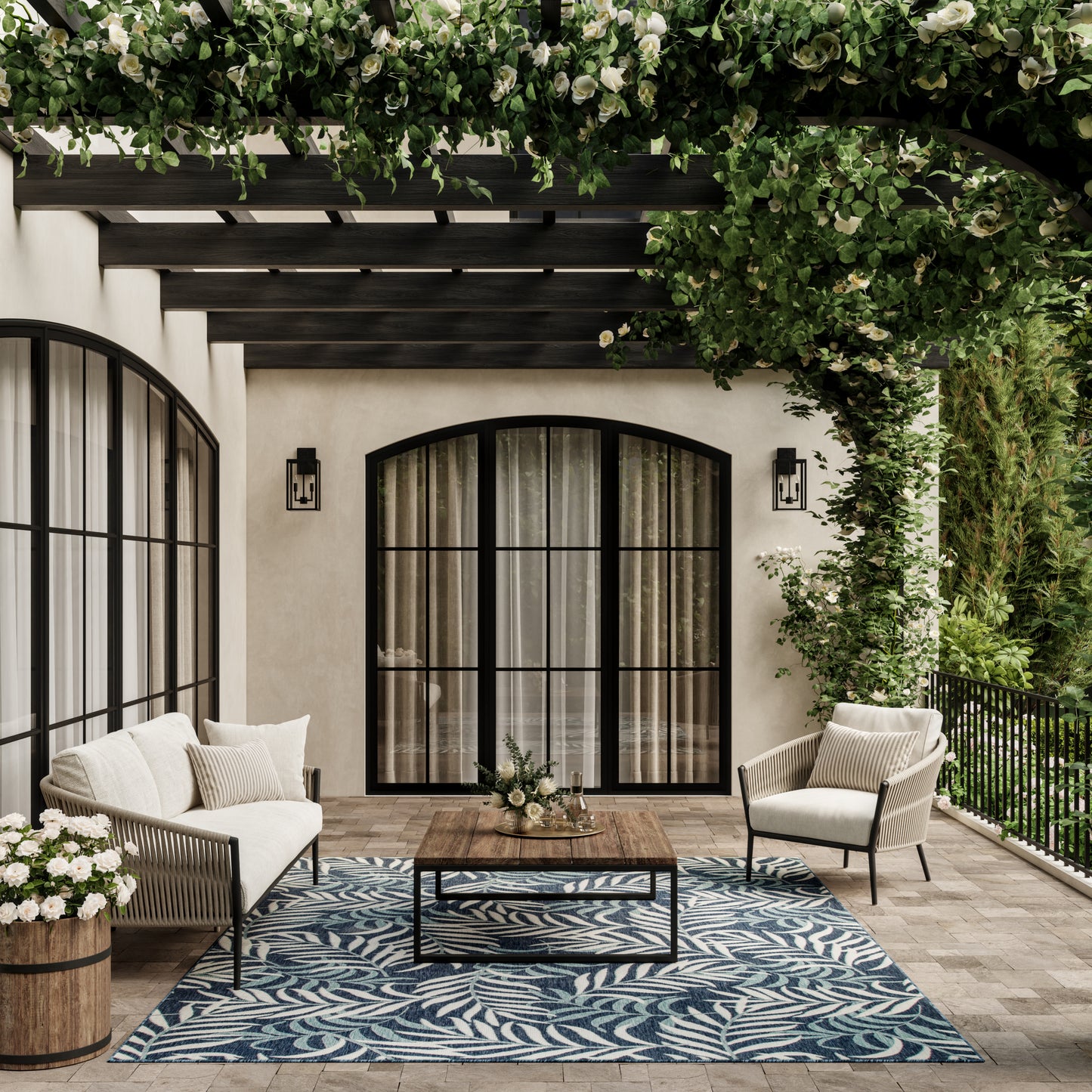 Elegant patio with beige cushioned seating, arched black-framed glass doors, blue leaf-pattern rug, and greenery.