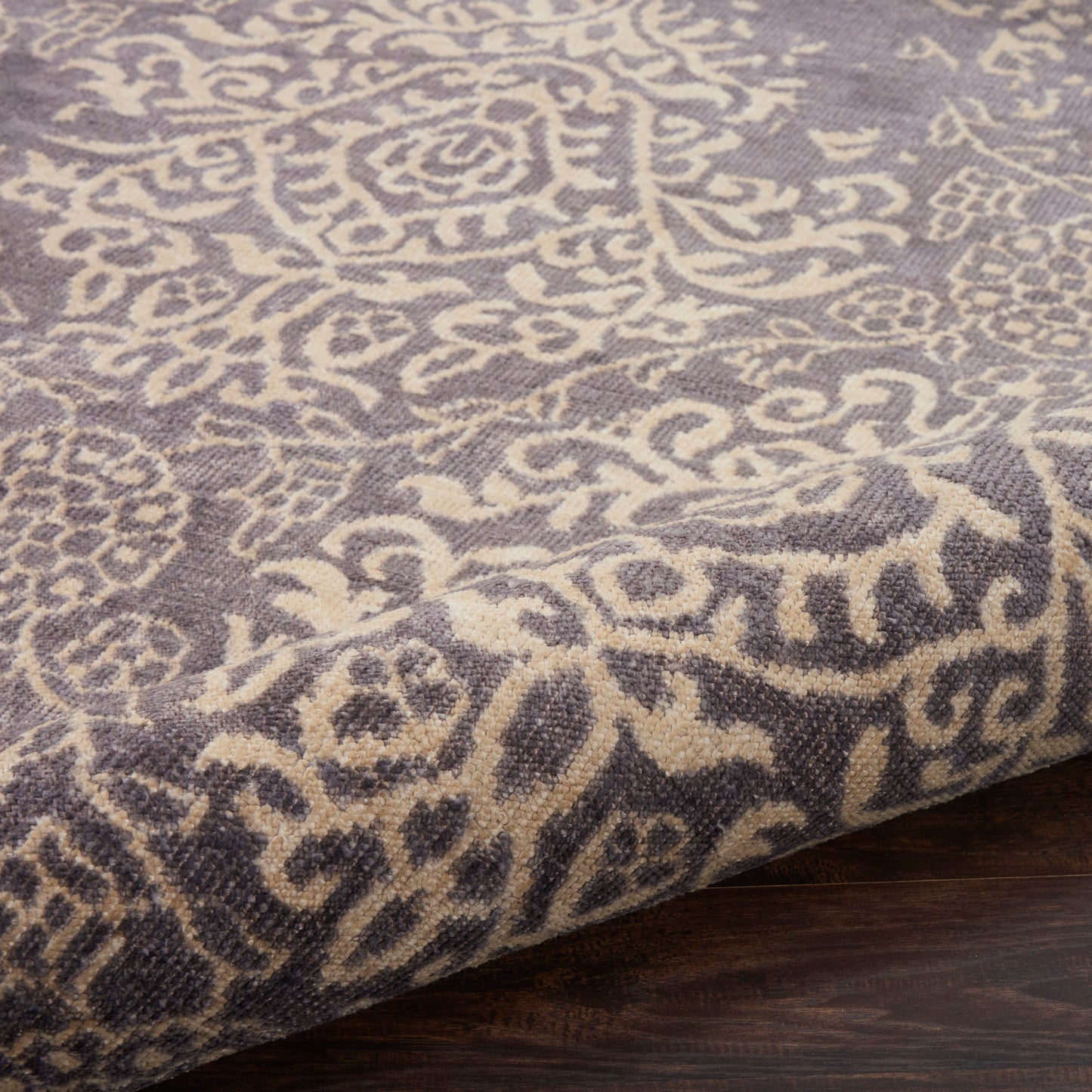 Grey-lavender rug with ivory damask pattern; woven texture, folded edge on dark wood floor.