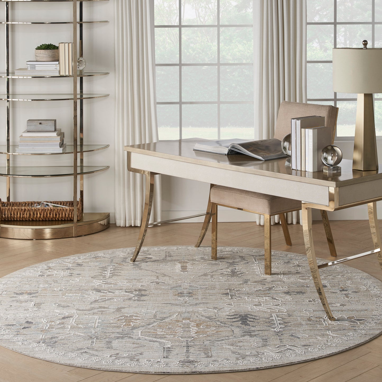 Modern home office with a round gray patterned rug, light-wood desk with gold legs, beige chair, gold lamp, and circular gold bookshelf beside a window.