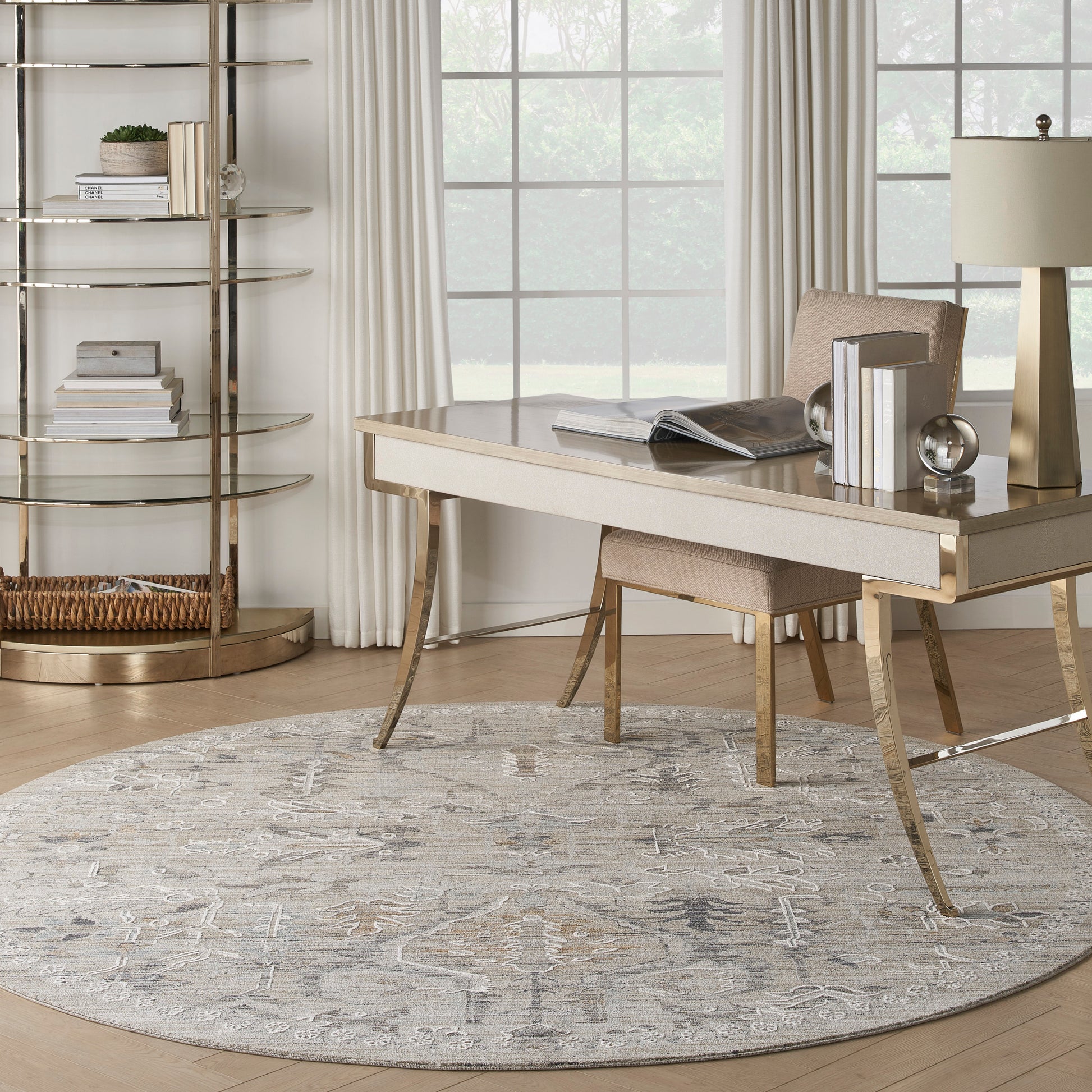 Modern home office with a round gray patterned rug, light-wood desk with gold legs, beige chair, gold lamp, and circular gold bookshelf beside a window.