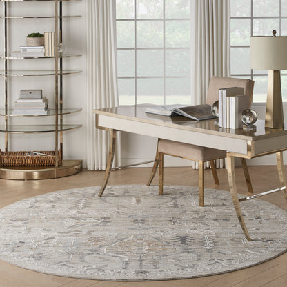 Modern home office with a round gray patterned rug, light-wood desk with gold legs, beige chair, gold lamp, and circular gold bookshelf beside a window.