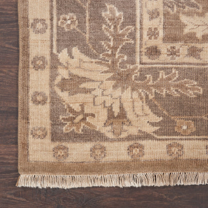 Beige-brown rectangular area rug with ornate floral border and fringe, laid on dark wood floor.