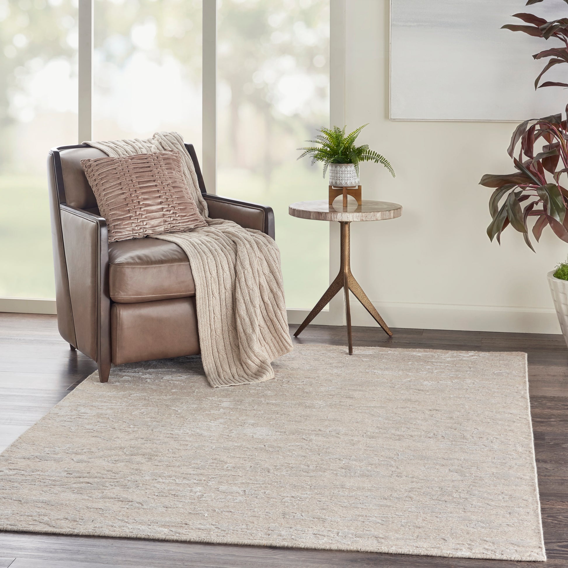 Beige area rug in a living room scene featuring a brown leather armchair, beige knitted throw, pillow, and a round wood side table.