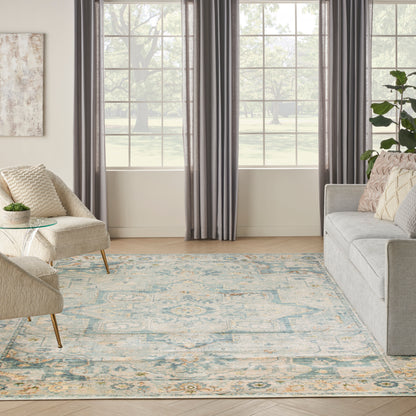 Living room with beige upholstered chair, light-blue patterned rug, gray sofa, dark gray curtains, glass side table and plants.