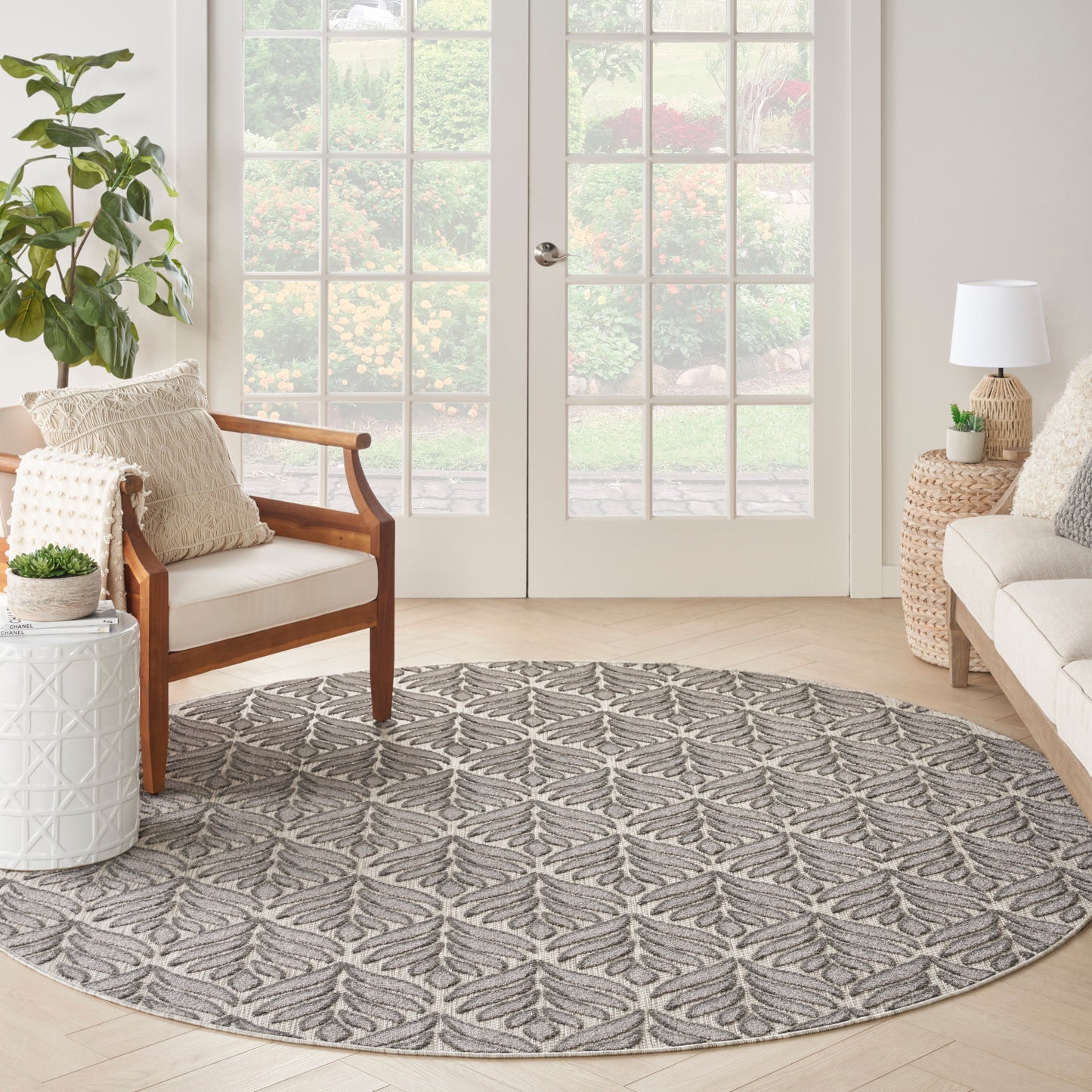 Grey round area rug with leaf motif in modern living room; wood armchair, wicker tables, beige sofa, glass French doors.