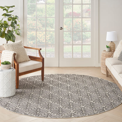 Grey round area rug with leaf motif in modern living room; wood armchair, wicker tables, beige sofa, glass French doors.