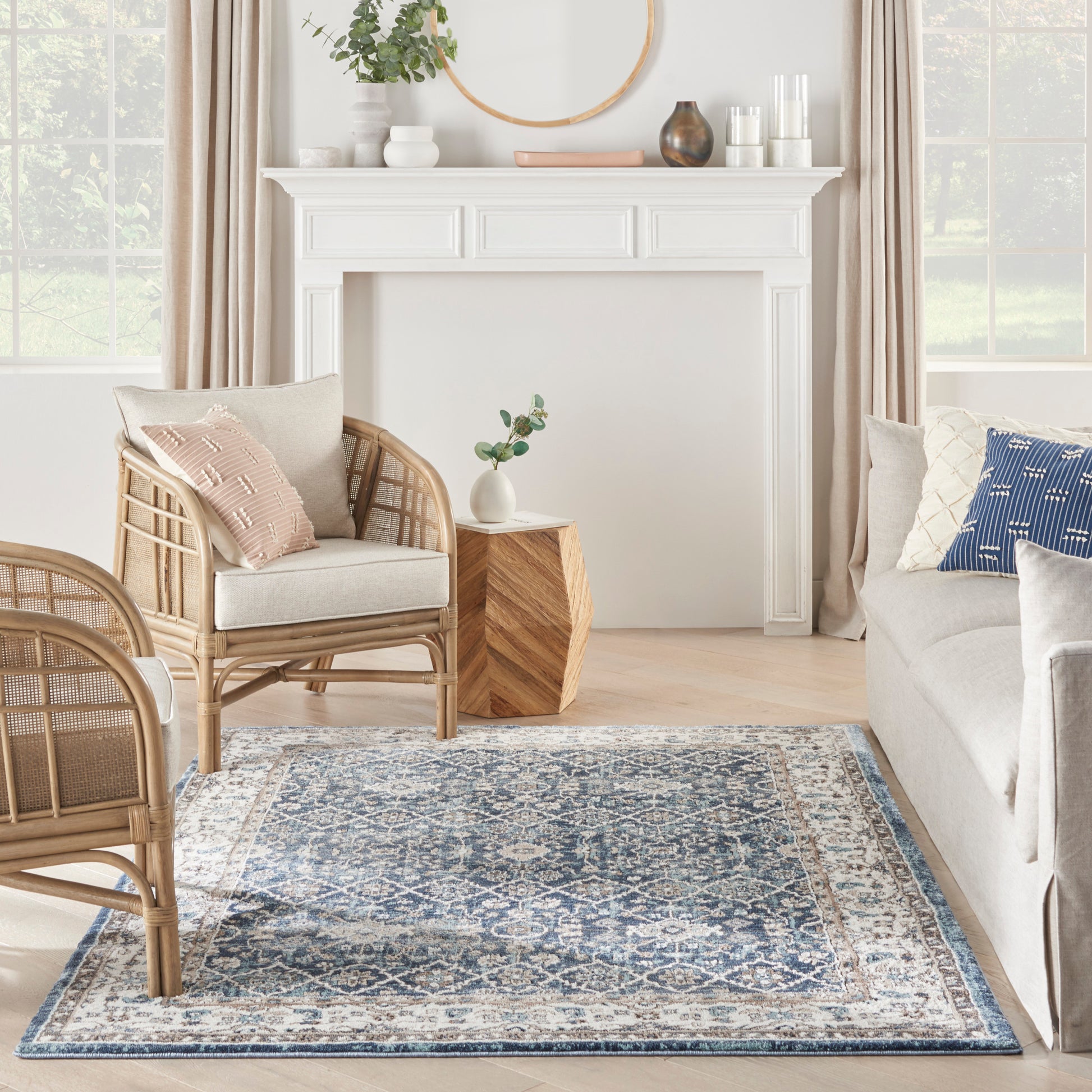 Bright living room with white mantel, round mirror, beige curtains, rattan chairs, blue patterned rug, and light sofa.