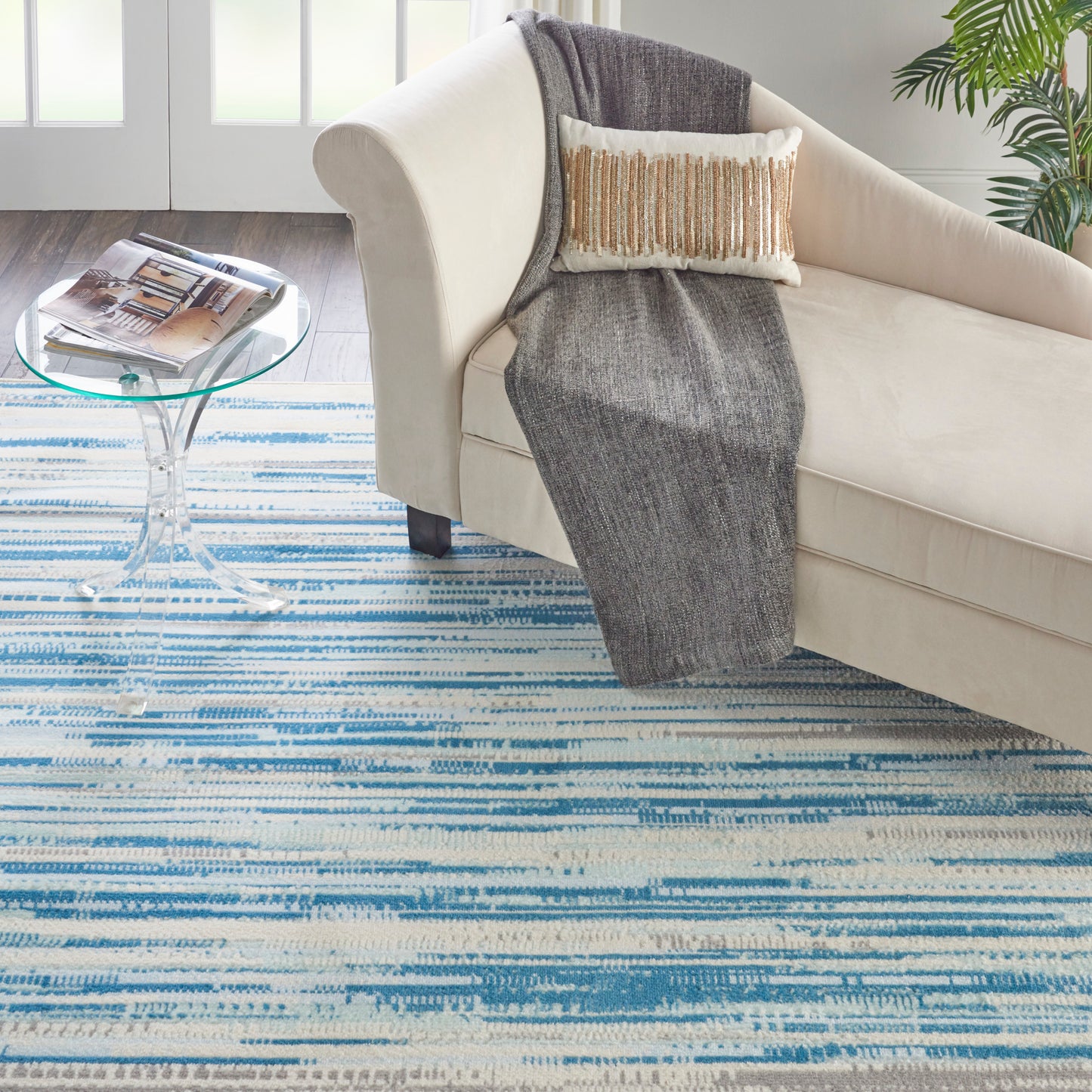 Cream sofa with gray throw and fringed pillow, round glass coffee table, blue striped rug, leafy plant in modern living room.
