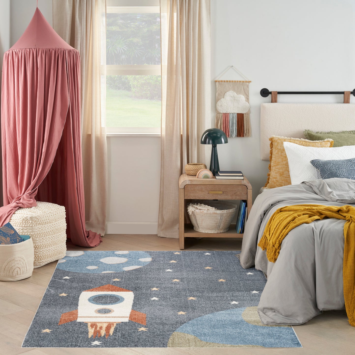Cozy kids' bedroom with pink canopy, blue space rocket rug, gray bedding, mustard throw, and light wood nightstand.