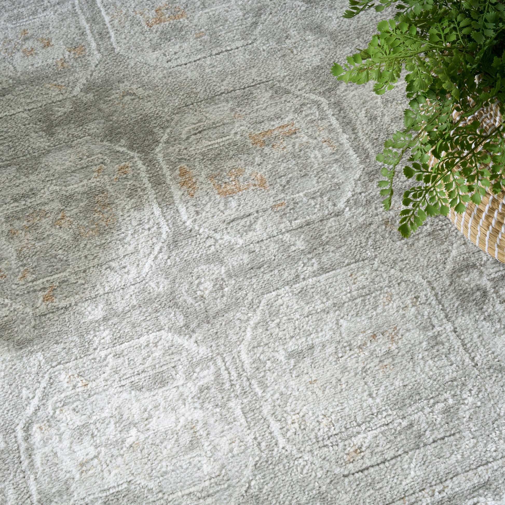 Light gray geometric patterned rug with soft pile and subtle tan accents; plant in a wicker basket nearby.