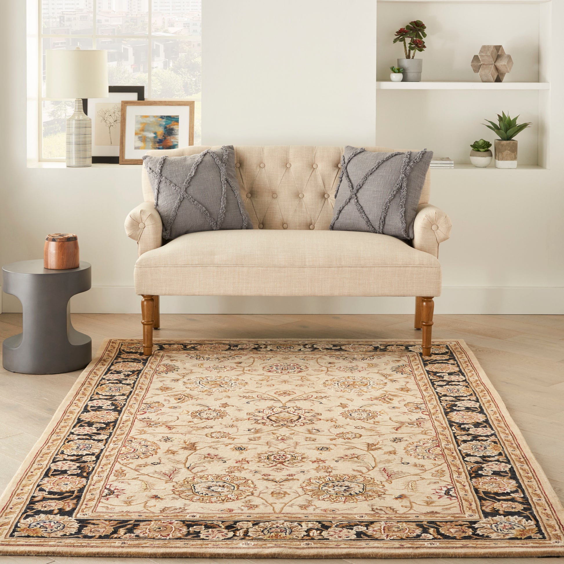 Beige tufted loveseat with gray textured pillows on an ornate beige area rug in a bright living room.