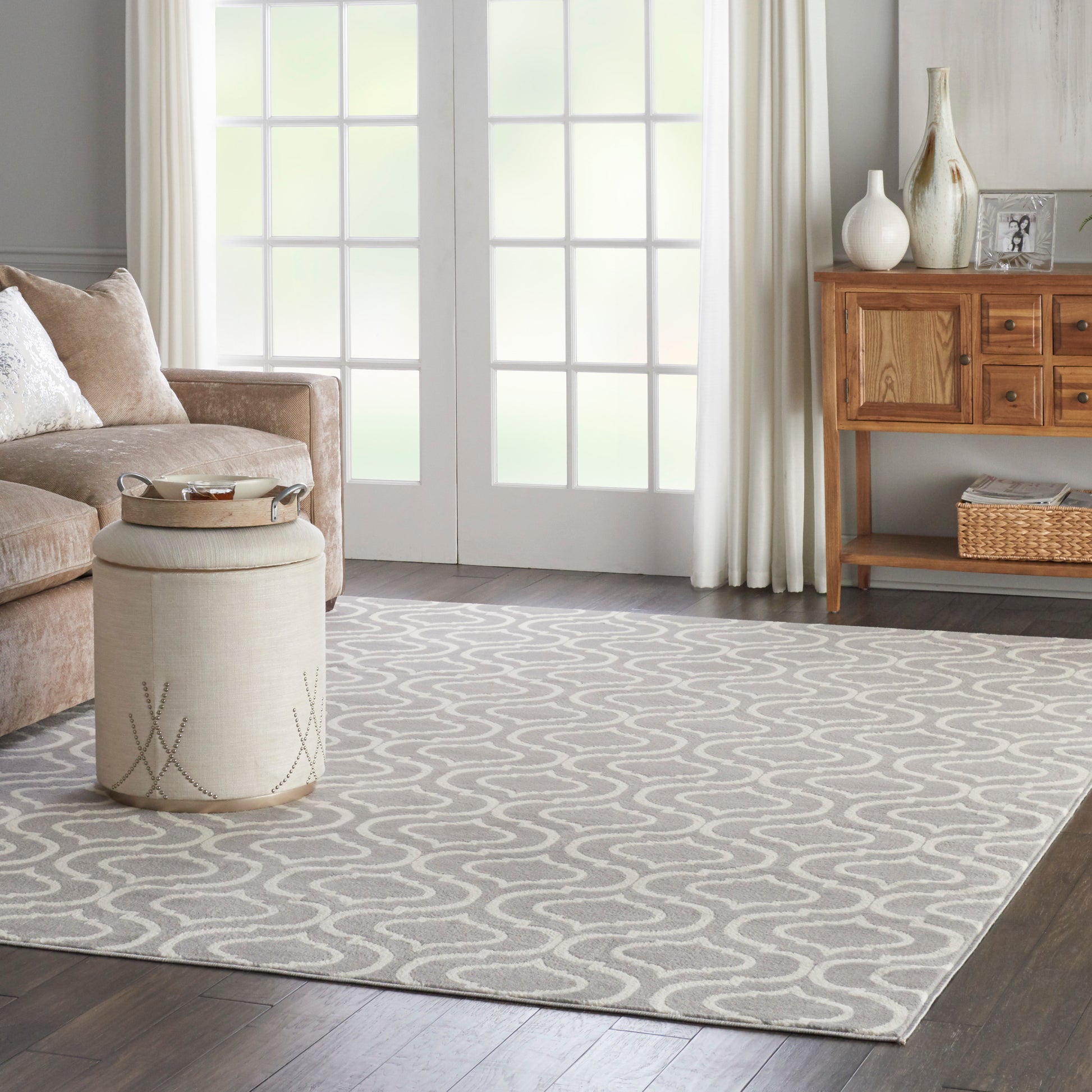 Light gray rug with cream wavy pattern on dark wood floor, beige sofa, round pouf, and wooden console.