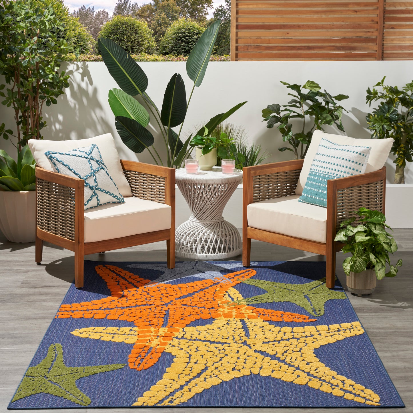 Outdoor rug with orange, yellow and green starfish on navy blue; wooden chairs, white cushions, wicker table.