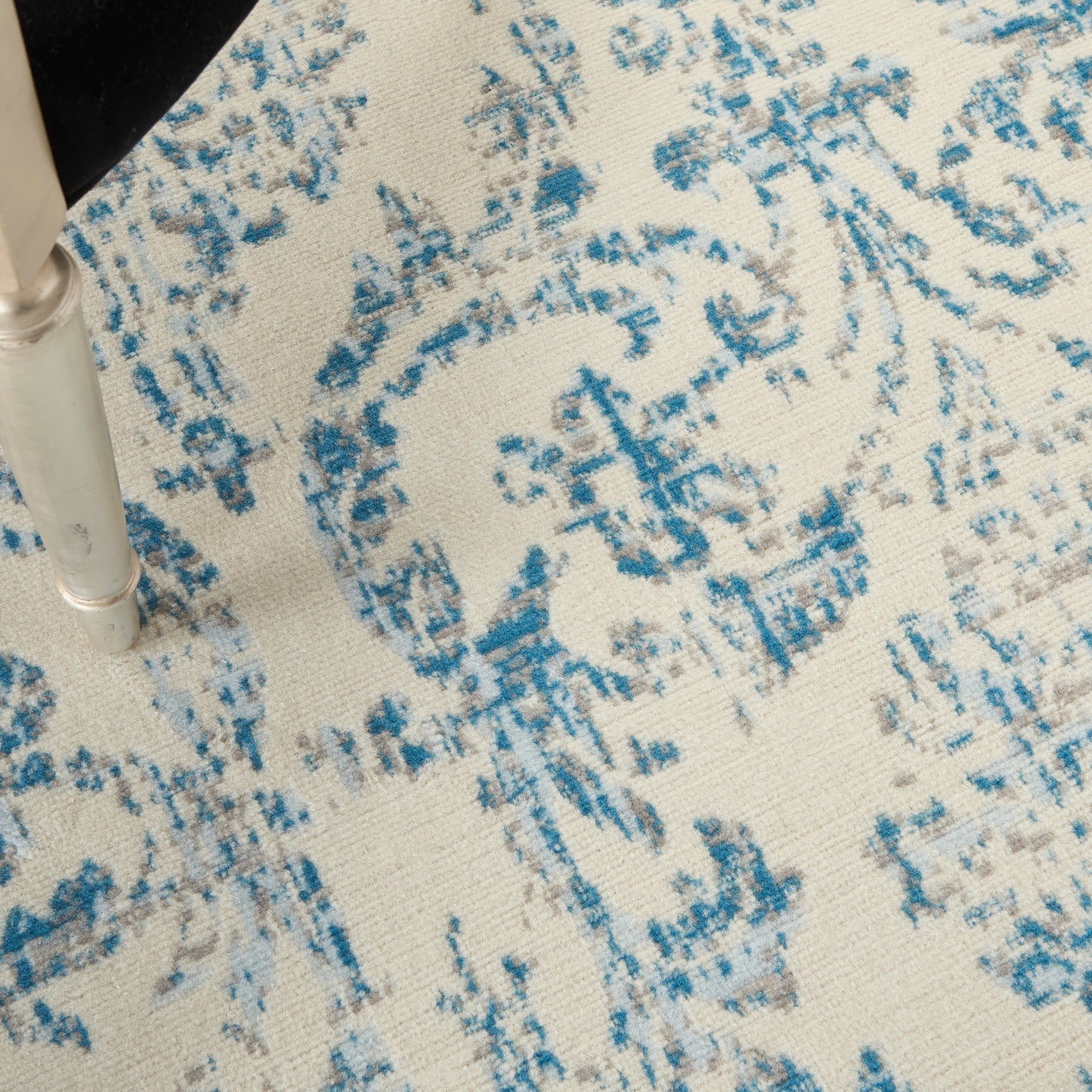 Beige rug with blue damask-style floral pattern; metal chair leg visible.