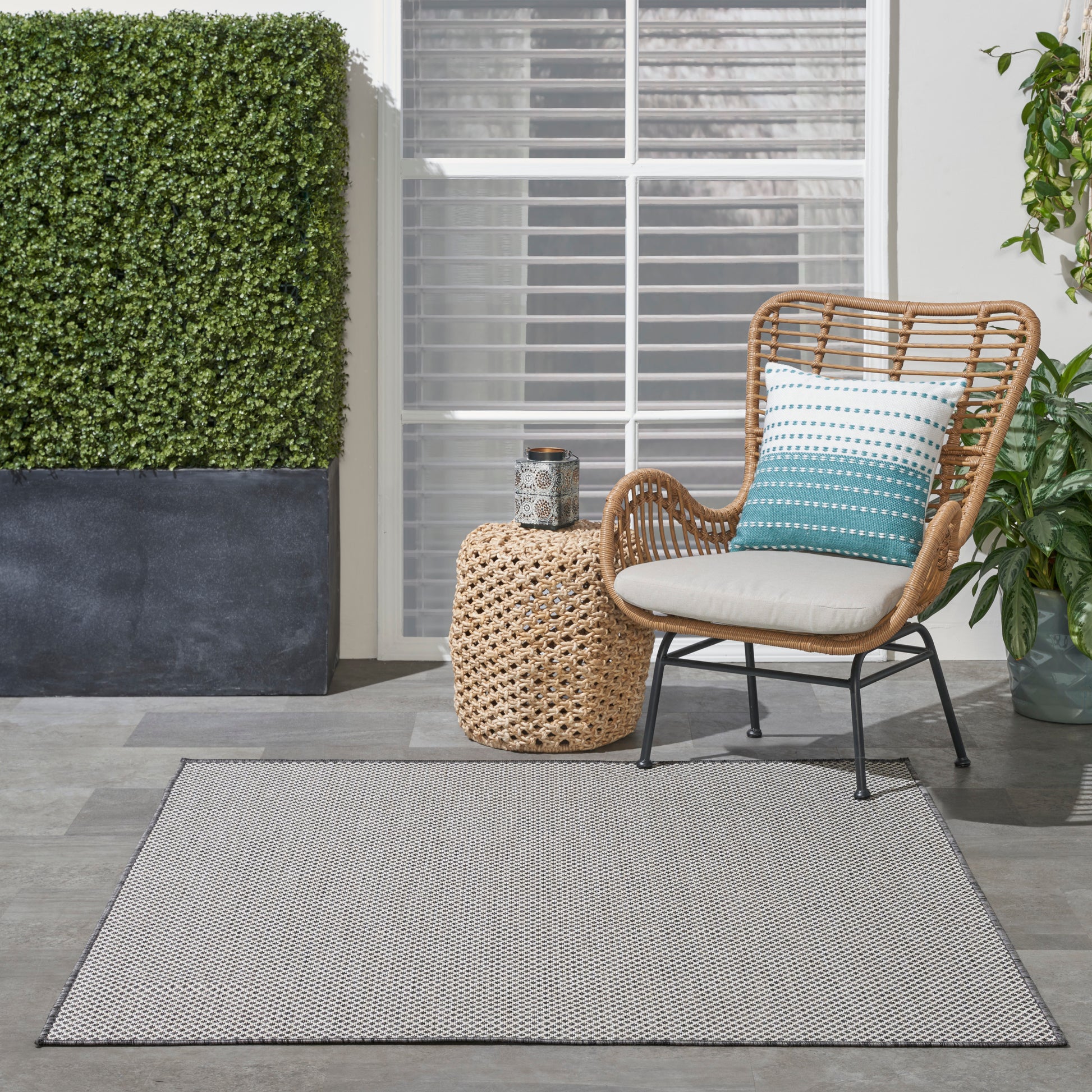 Outdoor patio: brown rattan chair with white cushion & teal patterned pillow, woven side table, gray rug, greenery.