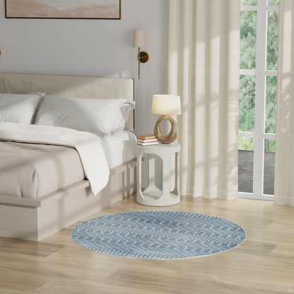 Round blue patterned rug beside a cream upholstered bed with white linens and a white side table in a bright bedroom.