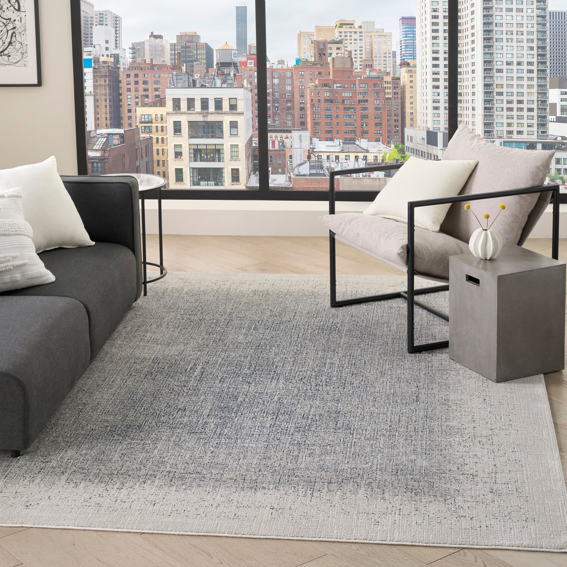 Modern living room with city view; grey sofa, light gray rug, black metal armchair, concrete side table with white vase.