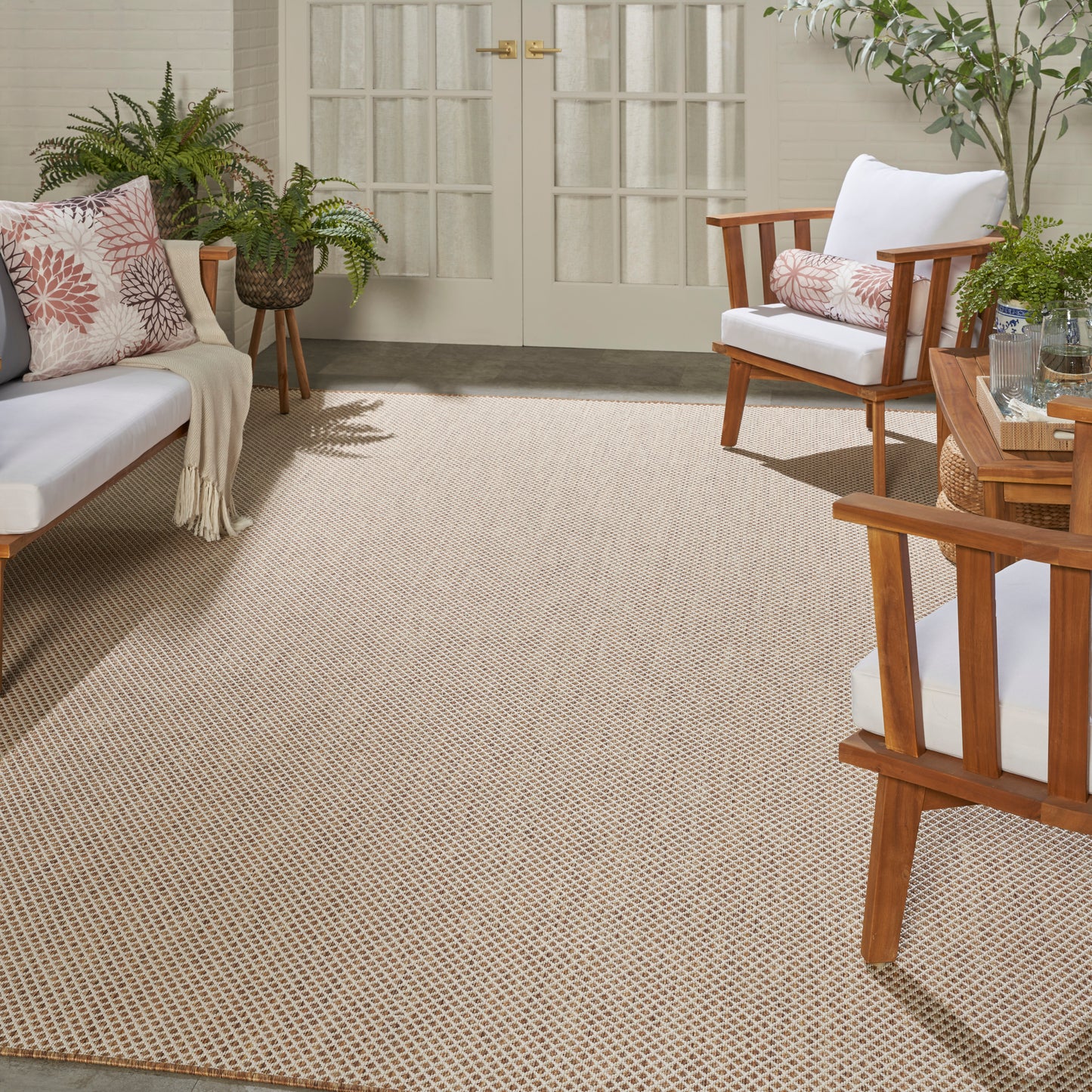 Sunlit porch with beige woven rug, mid-century wooden furniture, white cushions, pink-patterned pillows, and potted plants.