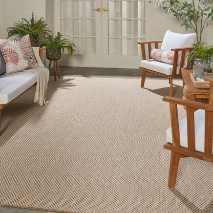 Sunlit porch with beige woven rug, mid-century wooden furniture, white cushions, pink-patterned pillows, and potted plants.