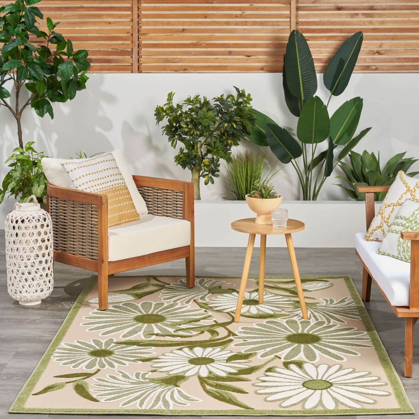 Outdoor floral rug on patio with wood-framed wicker armchairs, cream cushions & mustard pillows, round side table, & tropical plants.